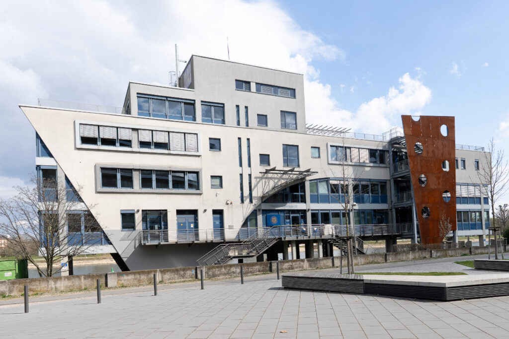 Außenansicht eines modernen, mehrstöckigen Bürogebäudes im Hafen Ludwigshafen mit markanter Architektur in Form eines Schiffsbugs. Das Gebäude liegt direkt an einer gepflasterten Uferpromenade am Wasser und repräsentiert hochwertige Gewerbeimmobilien im Hafenquartier.