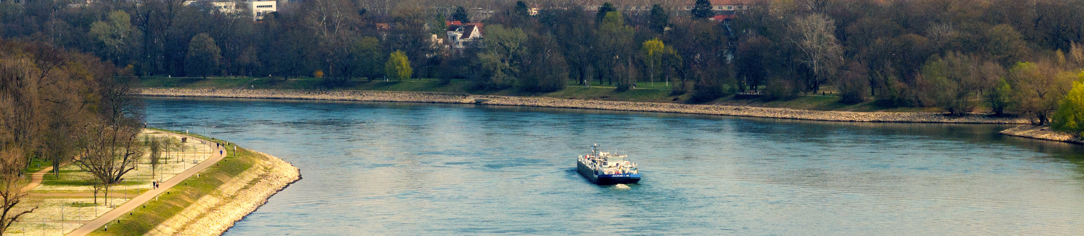 Aufnahme eines beladenen Binnenschiffs bei der Fahrt auf dem Rhein im Hafenbereich Ludwigshafen. Im Hintergrund sind industrielle Uferanlagen, Verladekräne und die Silhouette der Stadt unter blauem Himmel zu sehen.