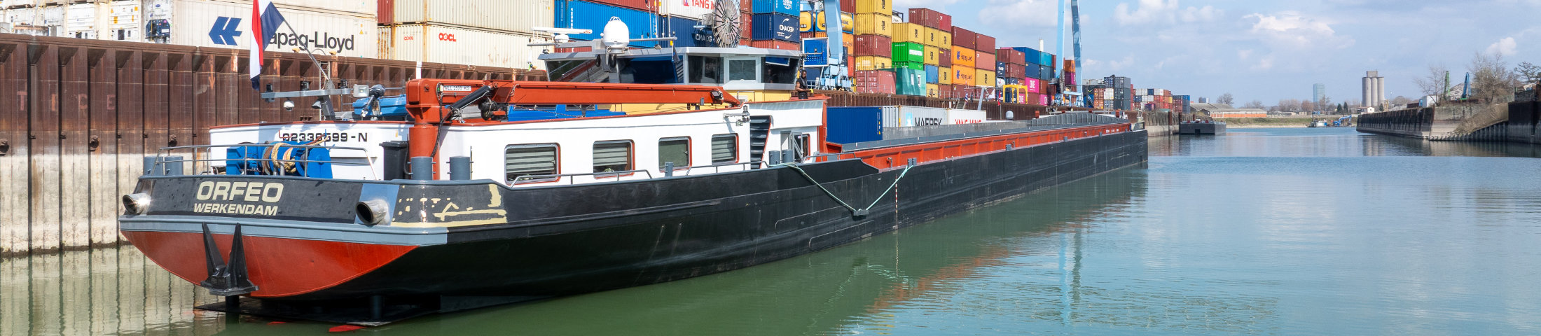 Binnencontainerschiff „Orfeo“ liegt im Hafenbecken vor gestapelten Frachtcontainern an einem Containerterminal.