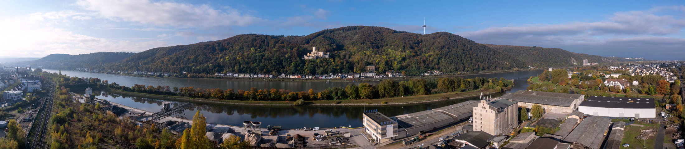 Panorama des Rheinhafens Lahnstein mit Industrieanlagen am Ufer; gegenüber der bewaldete Hang mit Schloss Stolzenfels über dem Rhein.