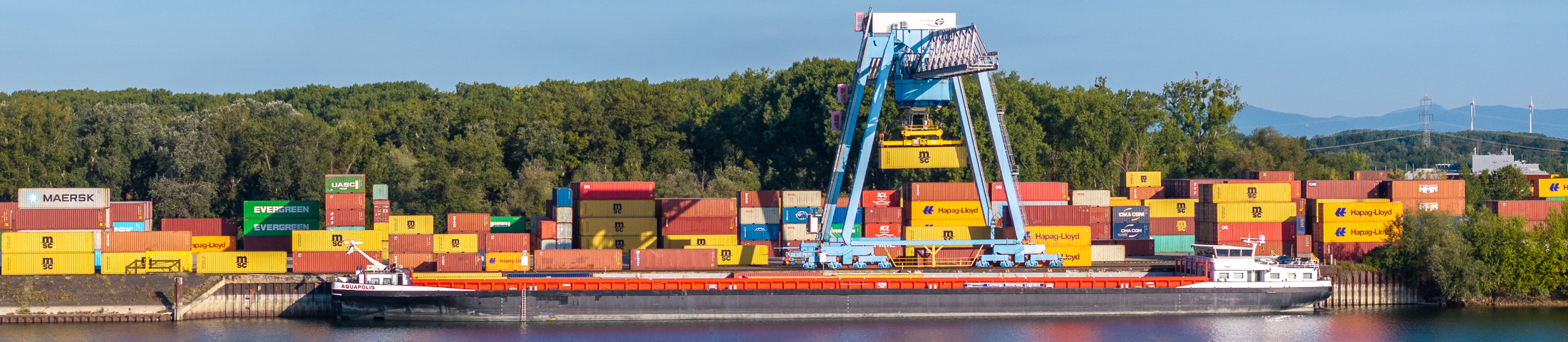 Containerschiff auf dem Rhein am Containerterminal Wörth; ein großer Portalkran verlädt Container vor gestapelten Boxen verschiedener Reedereien.