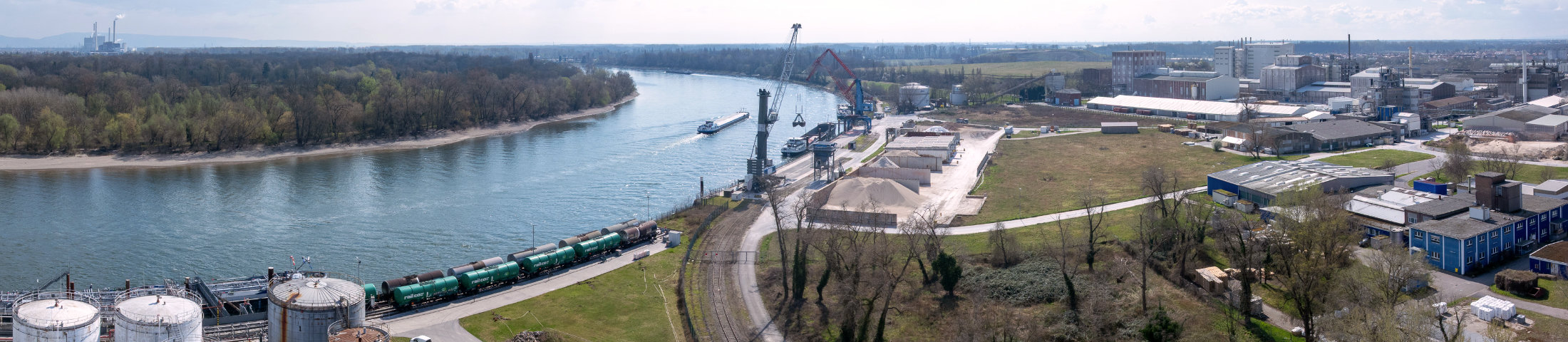 Panorama-Luftaufnahme eines Hafengeländes der Häfen Rheinland-Pfalz mit trimodaler Anbindung: Güterzug auf Gleisanlagen, Binnenschiff am Kai, Verladekräne und weitläufige Industrie- und Lagerflächen direkt am Rheinufer.